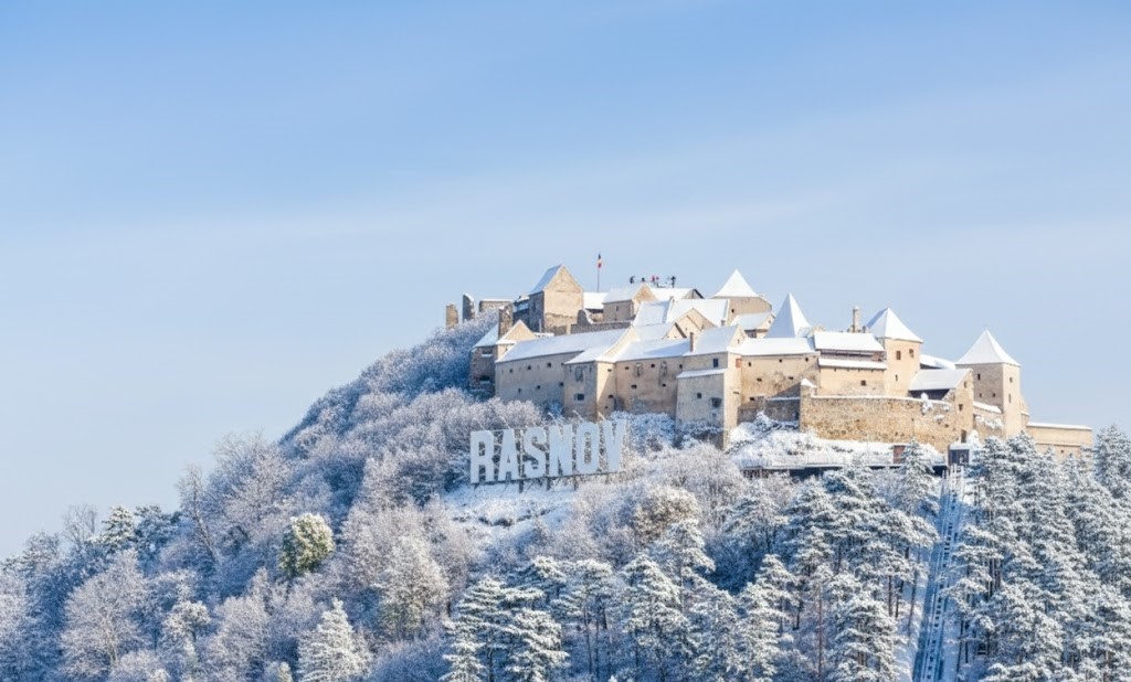 A realistic winter landscape of Râșnov Citadel in Romania. The ancient stone fortress sits atop a hill covered in snow-dusted pine trees. In the foreground, the large white 'RASNOV' Hollywood-style letters are prominent against the snowy slope, under a clear blue winter sky.