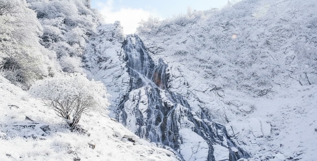 Wide shot of Capra Waterfall in winter with flowing water and snow-covered rocks in the Făgăraș Mountains.