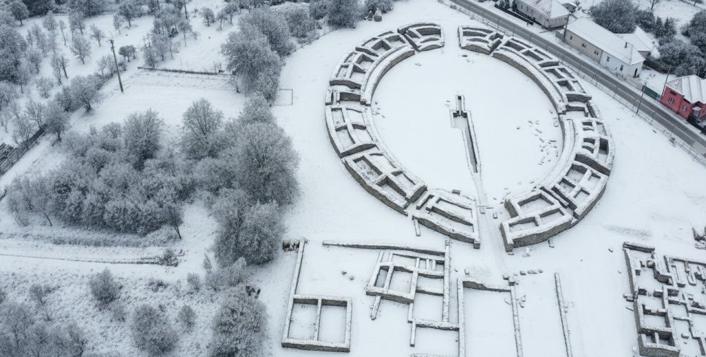 Ultra-wide aerial drone view of the Roman Amphitheater at Ulpia Traiana Sarmizegetusa during winter. The circular stone ruins and the surrounding archaeological site are covered in a fresh layer of white snow, with frost-covered trees and the nearby village visible in the cold, misty landscape.